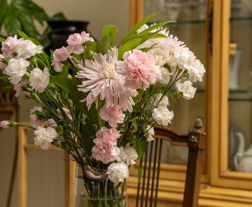 Pink and white mums and carnations in a vase in front of a china cabinet.