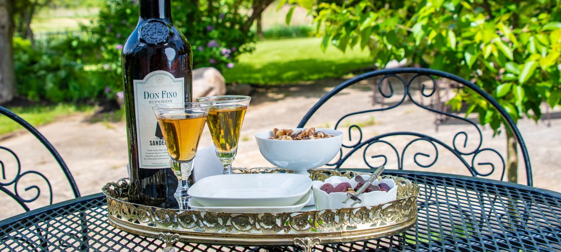 Bottle of liqueur and two glasses on a tray with nuts sits on a wrought-iron patio table. 